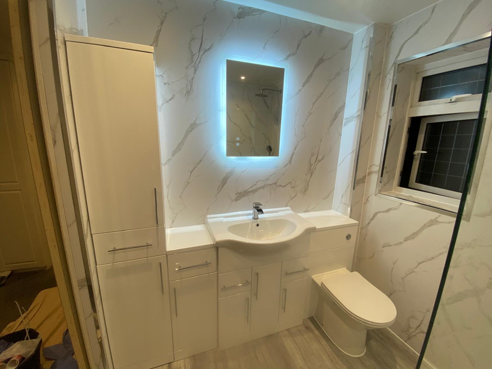 White vanity unit, WC and tall storage cupboard with an LED mirror on marble effect panelling