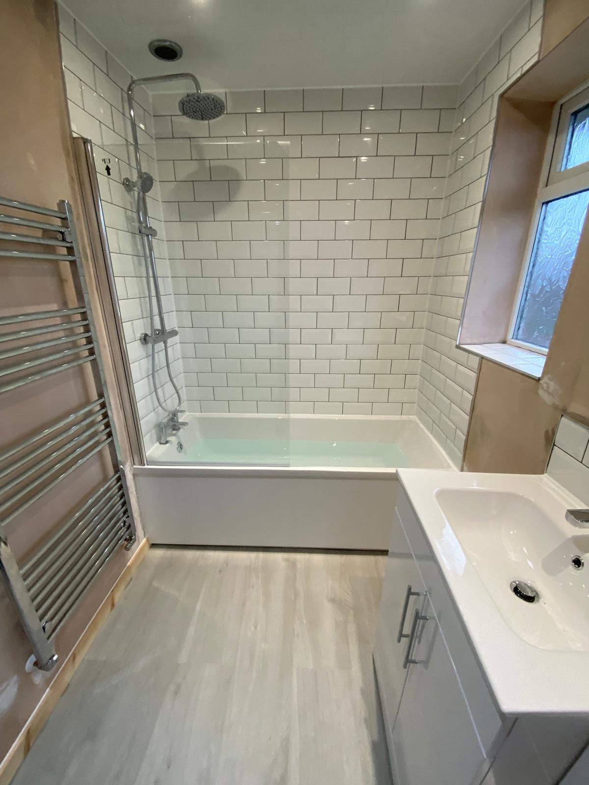 Finished bathroom with bath, shower over, white metro tiling, vanity unit and chrome towel rail