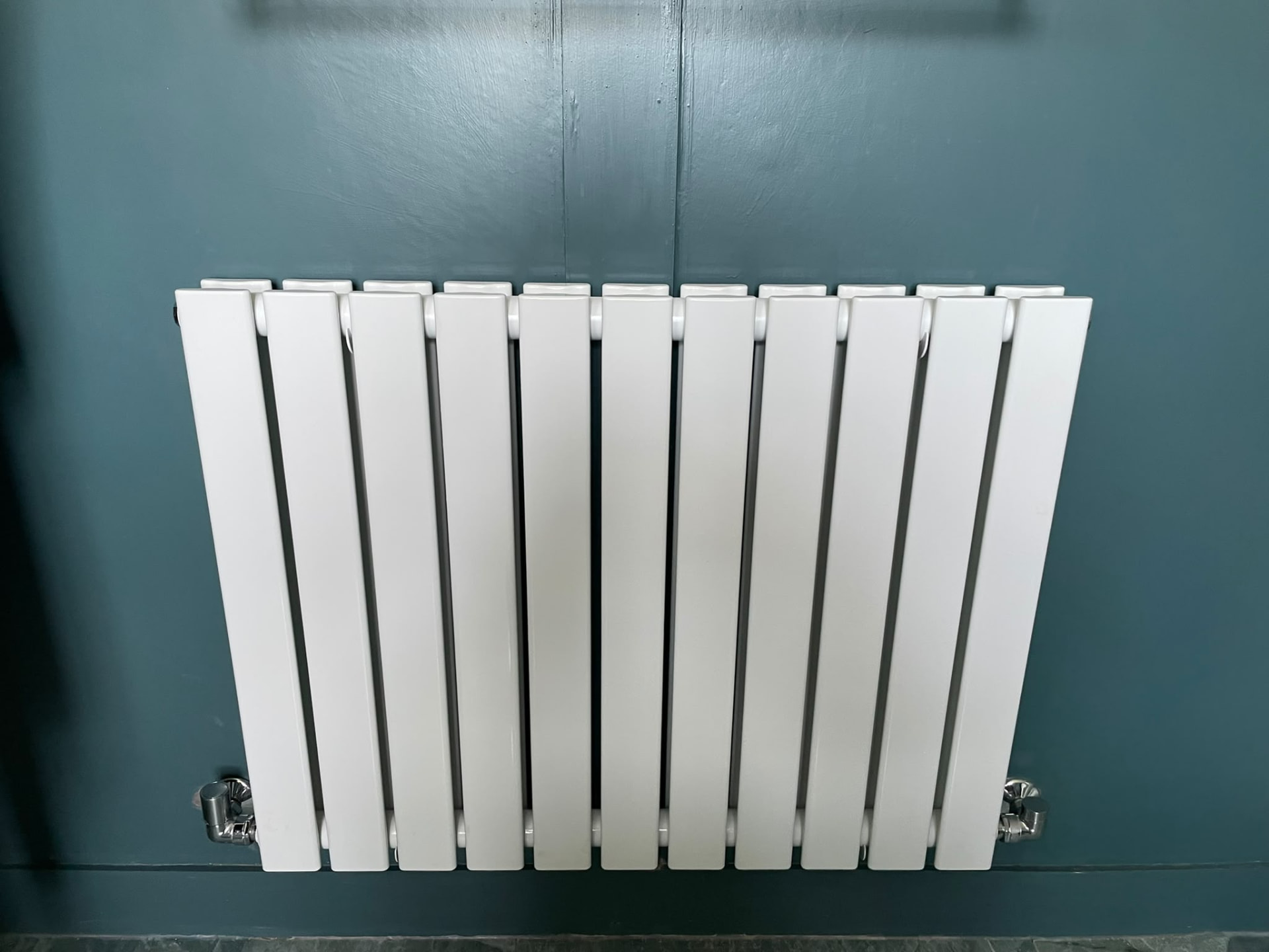 New white vertical designer radiator fitted on a dark teal wall, replacing an undersized radiator