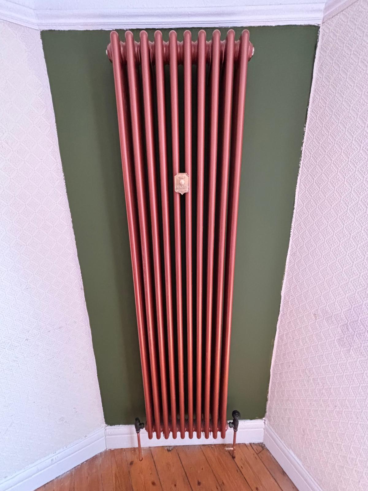 Tall copper coloured column radiator fitted in an alcove against a green wall